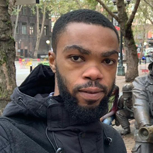 Picture of Bright Agbodzi, MS MPhil, in an outdoor plaza lined with trees, while another person interacts with a group of life‑sized firefighter statues arranged as if operating a fire hose