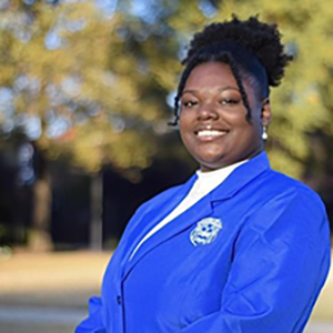 Picture of Shania Sanderson, M.S. standing outdoors in a natural, sunlit setting, wearing a bright royal‑blue blazer with an embroidered emblem on the chest and a white shirt underneath, posed with hands clasped in front against a softly blurred background of trees and grass.