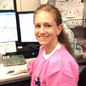 Picture of Tiffany R. Molinick, MPH, MLS(ASCP)CM wearing a bright pink lab coat and disposable gloves is seated at a workstation in a laboratory, working at a desk with multiple computer monitors and various lab supplies on shelving in the background.
